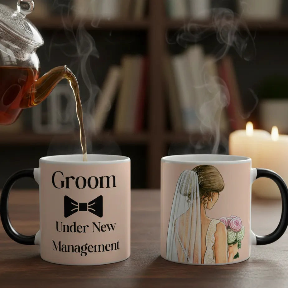 Tea being poured into a mug with 'Groom Under New Management' text, next to a mug with a bride illustration, on a wooden surface with books and a candle in the background.